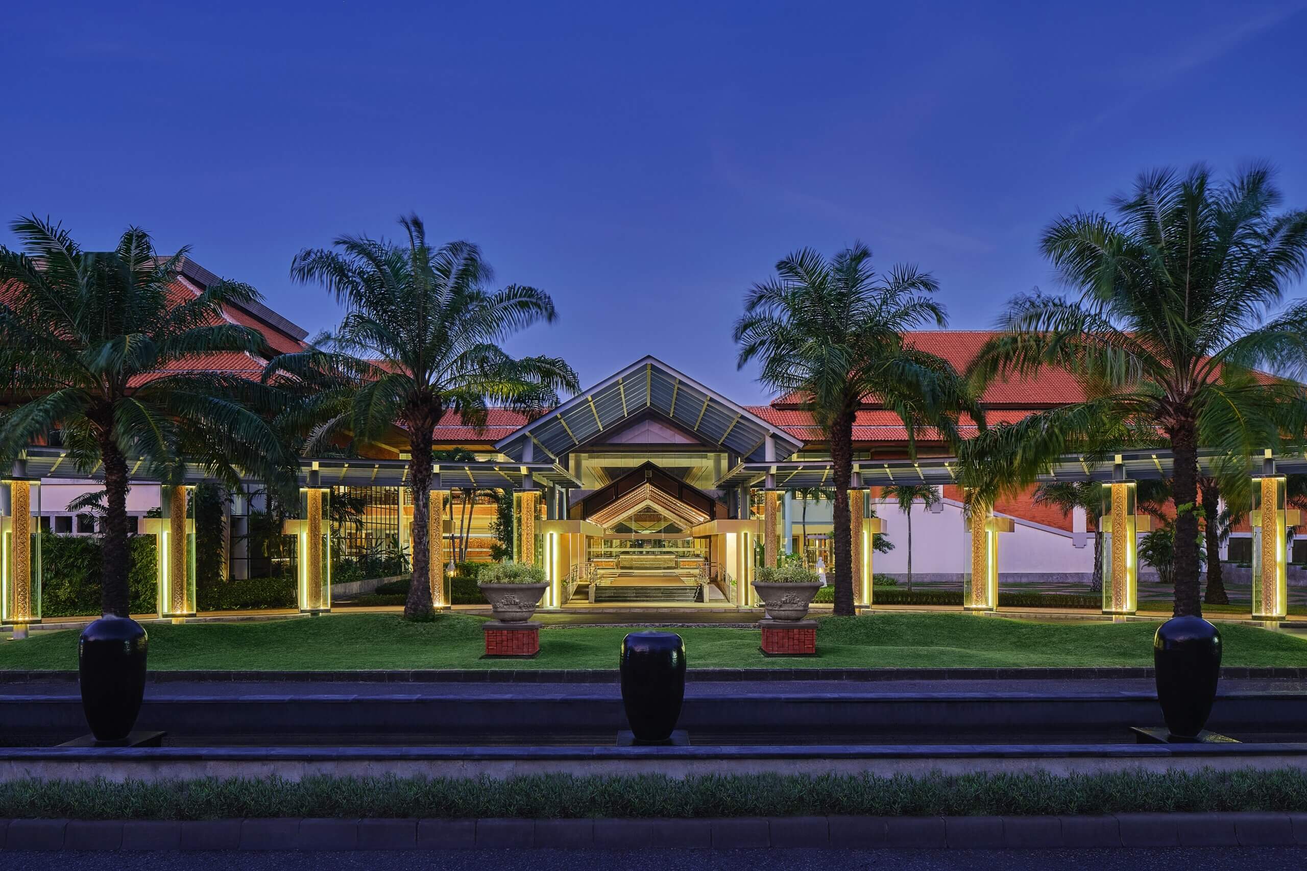 An evening view of the Bali International Convention Centre entrance, framed by palm trees and illuminated columns, with a red-roofed building in the background.