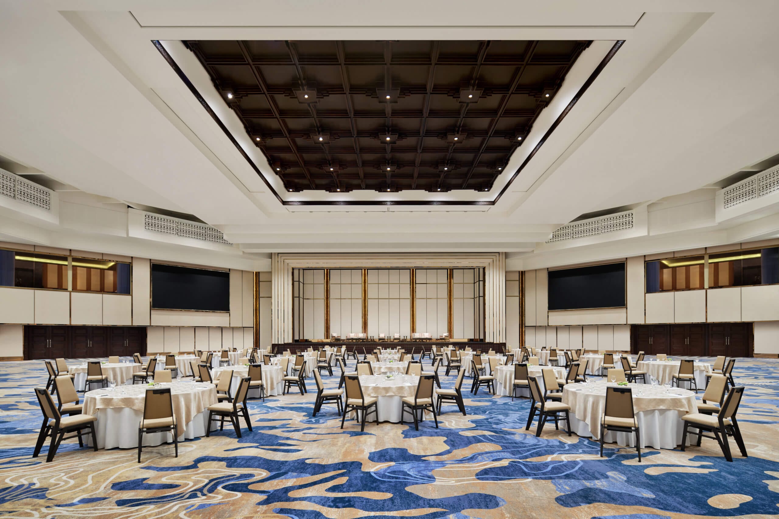 
A spacious, elegantly designed ballroom with round tables, cream-colored chairs, and a blue-patterned carpet, featuring a large stage and modern ceiling lighting.